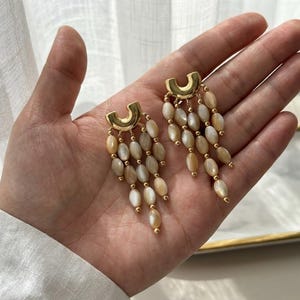 Peut inclure: Une paire de boucles d'oreilles dorées dotées d'une pièce supérieure en forme de U avec plusieurs brins pendants de perles ovales blanc cassé irisées. Les perles sont séparées par de petites sphères métalliques dorées, créant un design à franges.