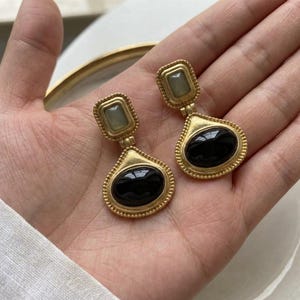 May include: A pair of gold-toned drop earrings featuring a rectangular top stud with a pale green stone and a larger, pear-shaped bottom pendant containing a smooth, polished black oval stone, both framed by a delicate beaded metal border.