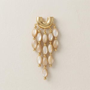 Può includere: Un ciondolo in metallo dorato caratterizzato da una parte superiore liscia a forma di U, da cui pendono cinque fili a cascata di perline ovali color bianco sporco iridescente. I fili sono intervallati da piccole sfere metalliche dorate.