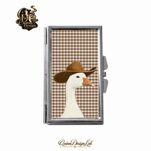 Può includere: Un astuccio rettangolare in metallo argentato con l'illustrazione di un'oca bianca che indossa un cappello da cowboy marrone su uno sfondo a quadretti vichy marroni e bianchi. L'astuccio è dotato di coperchio incernierato e finitura metallica.