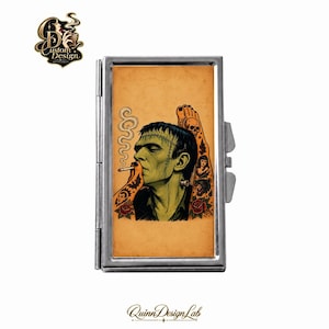May include: A rectangular silver metal cigarette case featuring a tan-colored insert with a green-skinned monster illustration. The artwork depicts a profile of a creature smoking, surrounded by tattoo-style imagery including a skull, roses, and faces.