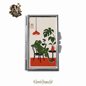 May include: A rectangular silver metal cigarette case featuring a graphic illustration of a green frog sitting on a black chair, drinking from a cup. The scene includes a red rug, a red pendant light, a potted monstera plant, and the red text characters 問一.