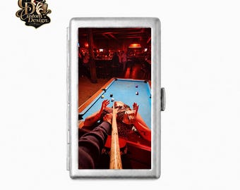 Könnte beinhalten: Ein rechteckiges Zigarettenetui aus silberfarbenem Metall mit einem aufgedruckten Bild einer Person, die in einer Bar Billard spielt. Das Bild zeigt ein Billard-Queue, das auf Kugeln auf einem blauen Tisch zielt, mit einer Person im Hintergrund. Der Text "Custom Design Available" und "QuinnDesignLab" sind zu sehen.