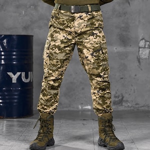 Op de afbeelding: Cargobroek met een digitaal camouflagepatroon in tinten olijfgroen, beige en zwart. De broek heeft zijzakken, een donker olijfgroene riem met een zwarte gesp en wordt gecombineerd met olijfgroene tactische veterschoenen.