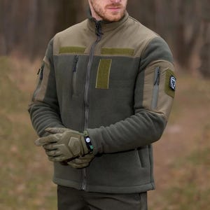 May include: An olive green tactical fleece jacket featuring a high collar, a full-length front zip, and multiple zipped pockets on the chest and sleeves. The jacket includes rectangular hook-and-loop panels on the chest and a small patch on the arm.