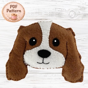 May include: A felt craft project featuring a brown and white dog face with long floppy ears, black eyes, and a black nose. A circular badge in the upper left corner displays the text PDF Pattern with an illustration of a spool of thread.