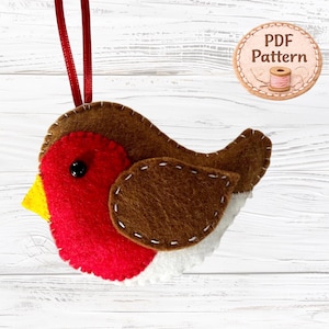 Può includere: Un ornamento in feltro a forma di pettirosso con testa e ala marroni, petto rosso brillante, becco giallo e ventre bianco. L'uccellino ha un piccolo occhio a perla nera ed è cucito con filo bianco. È presente un nastro rosso per appenderlo e un badge circolare con il testo PDF Pattern.