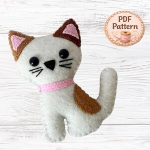 Può includere: Un peluche a forma di gatto in feltro bianco con punte delle orecchie marroni, un collare rosa e una macchia marrone sul fianco. Il gatto ha occhi a perla neri, un naso nero ricamato e baffi. Un badge circolare riporta il testo PDF Pattern.