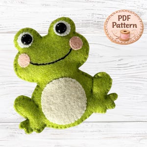 Könnte beinhalten: Eine grüne Frosch-Applikation aus Filz mit weißem Bauch, rosa Wangen und schwarzen Kunststoffaugen. Der Frosch hat ein aufgesticktes Lächeln und ein rundes Abzeichen mit dem Text PDF Pattern sowie einer Abbildung einer Garnrolle.