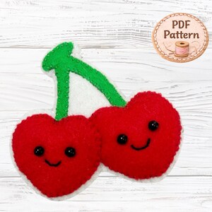May include: A felt craft project featuring two bright red heart-shaped cherries with black bead eyes and stitched smiles, connected by a green felt stem. A circular badge in the upper right corner displays the text PDF Pattern with a spool icon.