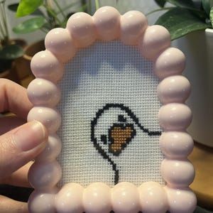 Op de afbeelding: Een kleine, boogvormige kruissteekborduurwerk met een zwart omlijnde ganzenkop met een oranje snavel op een witte stoffen achtergrond. Het werk is gevat in een glanzende, lichtroze lijst met een afgeronde, bubbelachtige textuur.