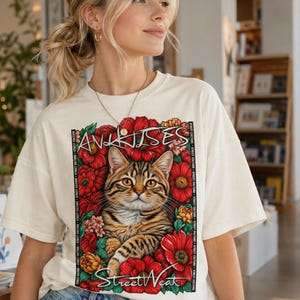 May include: A cream-colored short-sleeve t-shirt featuring a graphic print of a brown tabby cat surrounded by vibrant red and orange flowers. The text "ANKHISES" appears above the cat, and "Street Weak" is printed below, framed by vertical borders.