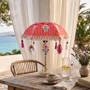 Puede incluir: Una lámpara de mesa con una pantalla de tela a rayas de color naranja y rosa vibrante con borde festoneado, flecos de color crema y borlas decorativas colgantes. La lámpara tiene una base y un soporte sencillos de color crema.