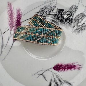 Puede incluir: Una pulsera de cuentas tejidas con un patrón geométrico de pequeñas cuentas cuadradas de color turquesa y oro metálico. La pulsera está construida con un diseño de cuadrícula repetitivo, mostrando una disposición consistente de los colores.