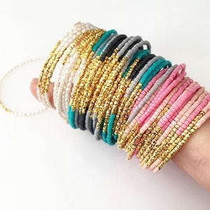 Könnte beinhalten: Ein Stapel bunter Perlenarmbänder mit goldenen Akzenten. Die Armbänder bestehen aus kleinen, runden Perlen in den Farben Rosa, Grau, Türkis und Weiß. Einige der Armbänder haben ein goldenes Band, das entlang der Kante verläuft.