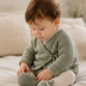 May include: A sage green knitted wrap cardigan with long sleeves and matching knitted booties. The cardigan features a delicate openwork pattern, ribbed cuffs, and a side tie closure. The booties are also knitted in the same sage green yarn.