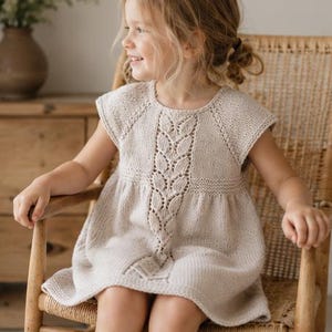 Puede incluir: Un vestido de punto beige que presenta un diseño central vertical de cable con patrón de hojas, un cuello redondo y mangas cortas. La prenda está confeccionada con un punto texturizado y un dobladillo liso, mostrando una construcción clásica.