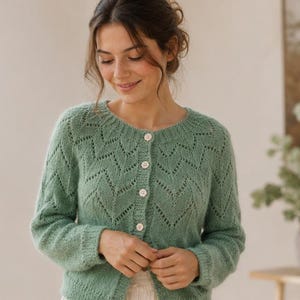 Könnte beinhalten: Eine salbeigrüne Strickjacke mit einem zarten Chevron-Spitzenmuster, einem Rundhalsausschnitt, langen Ärmeln mit gerippten Bündchen und einer Knopfleiste auf der Vorderseite mit vier kleinen, hellen, runden Knöpfen. Das Kleidungsstück hat eine weiche Textur.