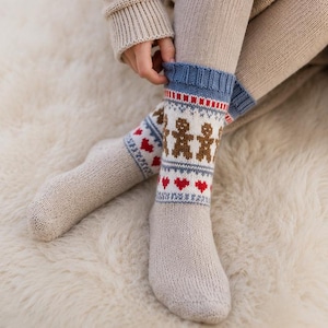 May include: Beige knit socks featuring a blue ribbed cuff and a patterned band with brown gingerbread men and red hearts. The socks are crafted from a textured yarn and extend to the mid-calf, showcasing a detailed fair isle style design.