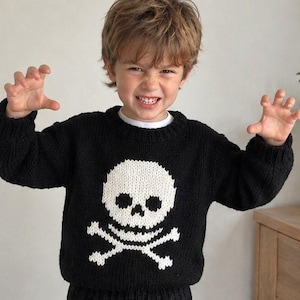 May include: A black long-sleeved knit sweater featuring a prominent white skull and crossbones graphic on the front. The sweater has a ribbed crew neckline, ribbed cuffs, and a ribbed hem, constructed with a thick, chunky yarn texture.