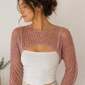 May include: A dusty rose long-sleeve crochet shrug featuring an open-knit mesh pattern with a wide rounded neckline, worn over a plain white sleeveless top and light-colored pants. The shrug has a cropped silhouette with long sleeves.