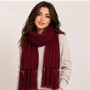 May include: A deep burgundy knitted scarf featuring a thick, ribbed texture and long, straight fringe detailing along the bottom edges. The scarf is wrapped around the neck and hangs down over a light beige, long-sleeved, textured knit sweater.