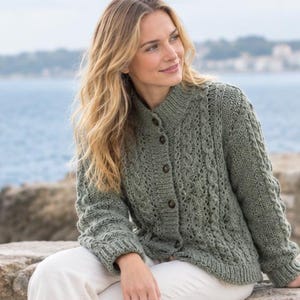 May include: A sage green cable-knit cardigan featuring a high collar, long sleeves with ribbed cuffs, and a front button closure with dark brown buttons. The knit pattern includes vertical cable textures and intricate stitch details throughout.