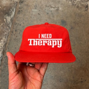 Pode incluir: Um boné de beisebol vermelho brilhante com o texto branco I NEED THERAPY impresso no painel frontal. O boné tem uma copa estruturada, uma aba reta e um botão no topo. O design está centralizado e utiliza uma fonte em negrito sem serifa.