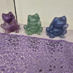 May include: Three small frog figurines in purple, green, and dark blue, each depicted in a seated position with one hand raised. The frogs have a smooth, semi-translucent finish and are arranged in a row on a flat surface.