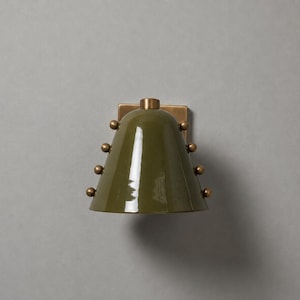 May include: A wall sconce featuring a glossy olive green bell-shaped shade. The shade is accented with a vertical row of three small brass spheres on each side. A square brass mounting plate sits behind the top of the shade, connecting it to the wall.