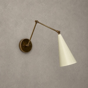 Può includere: Una lampada da parete con braccio orientabile dotata di una base circolare in ottone anticato e un braccio in metallo coordinato. L'apparecchio include un paralume conico color bianco sporco che dirige la luce verso il basso, con giunti regolabili.