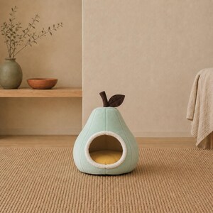 May include: A pear-shaped pet bed in a soft mint green color with a dark brown stem and leaf on top. The entrance is circular with a white fuzzy trim, and the interior floor of the bed is a bright yellow cushion, creating a cozy space for pets.