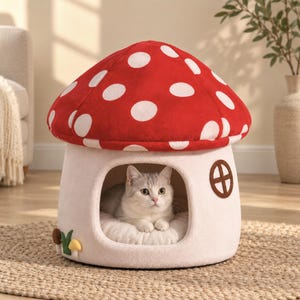 Puede incluir: Una cama para mascotas con forma de casa de hongo con un techo rojo de lunares blancos. La base es de color crema con una ventana circular y una pequeña entrada arqueada donde un gato de color claro descansa sobre un cojín blanco suave.