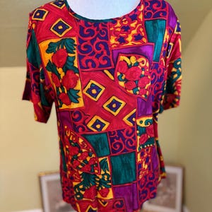 May include: A short-sleeved blouse featuring a vibrant, multi-coloured patchwork pattern. The design includes geometric blocks and floral motifs in bold shades of red, purple, yellow, and green, with a round neckline and a relaxed fit.