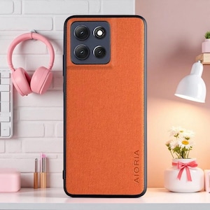 May include: A rectangular smartphone case with a textured orange back panel and a black frame. The camera module is square with rounded corners, containing three lenses and a flash. The brand name AIORIA is embossed vertically on the lower right.