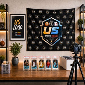 May include: A black wall tapestry featuring a repeating US logo pattern and a central emblem with a basketball, football, and baseball. Below, five graded sports trading cards are displayed on a wooden desk next to a camera on a tripod.