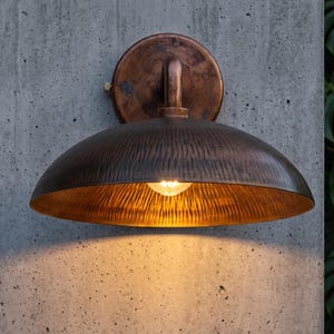 May include: A wall-mounted light fixture featuring a circular backplate and a wide, dome-shaped shade with a textured, hammered metal finish in a dark bronze tone. The lamp emits a warm, glowing light from a single bulb centered underneath.