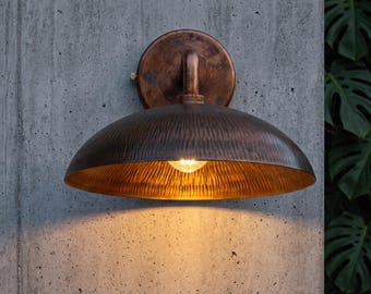 May include: A wall-mounted light fixture featuring a circular backplate and a wide, dome-shaped shade with a textured, hammered metal finish in a dark bronze tone. The lamp emits a warm, glowing light from a single bulb centered underneath.