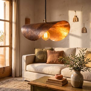 Könnte beinhalten: Eine Pendelleuchte mit einem breiten, geschwungenen, gehämmerten Kupferschirm mit warmem Metallic-Finish. Die Leuchte ist an einem schwarzen Kabel aufgehängt und enthält eine einzelne, freiliegende Edison-Glühbirne, die ein sanftes, goldenes Licht abgibt.