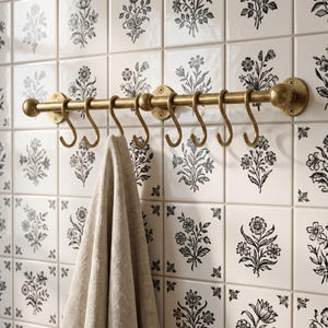 May include: A brass-toned metal rail mounted to a wall with three circular brackets, featuring eight matching S-shaped hooks hanging from the rod. A light beige linen-textured cloth hangs from one of the hooks, positioned against a tiled wall.