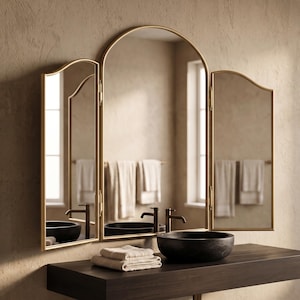 May include: A tri-fold vanity mirror with a thin gold metal frame. The central panel features an arched top, while the two side panels are hinged and adjustable. The mirror reflects a bathroom setting with a dark sink and white towels.