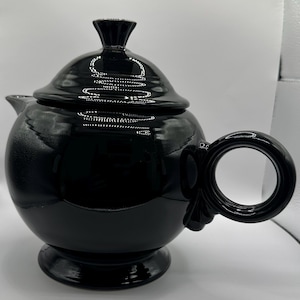 May include: A glossy black teapot with a spherical body, a matching lid with a small knob handle, a circular side handle, and a pedestal base. The surface is smooth and reflective, showing light highlights across the rounded exterior form.