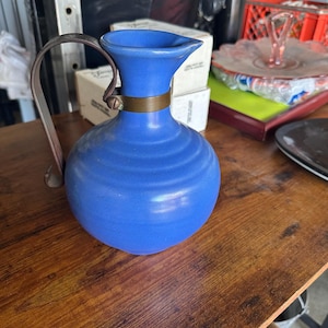 May include: A blue ceramic pitcher with a rounded body and horizontal ribbed texture. The vessel features a flared rim and a dark metal handle attached to the neck with a circular metal band secured by a single screw on the front side.
