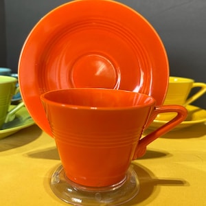 May include: A bright orange ceramic teacup and matching saucer set. The teacup features a smooth, glossy finish with subtle horizontal ring detailing around the body and a curved handle. The saucer is round with concentric circular ridges.