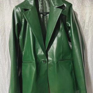 Könnte beinhalten: Ein waldgrüner Blazer mit glatter, glänzender Oberfläche. Diese langärmelige Jacke verfügt über einen fallenden Reverskragen, einen einzelnen Knopfverschluss auf der Vorderseite und zwei horizontale Leistentaschen an den unteren Vorderteilen.