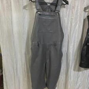 May include: A pair of charcoal gray overalls featuring a front bib pocket with a horizontal zipper closure, adjustable shoulder straps with metal buckles, and side waist buttons. The garment has a straight-leg cut and a smooth, matte finish.