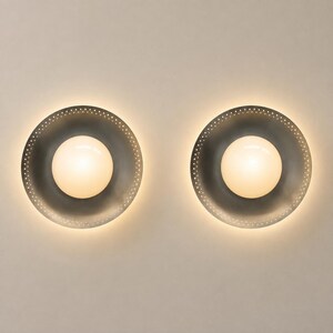 May include: Two circular wall sconces featuring a dark gray metal shade with a perforated edge and a central glowing white glass globe. The lights emit a soft, warm illumination, creating a halo effect around the circular metal fixture.