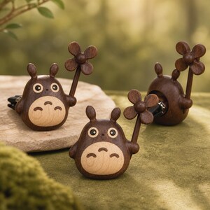 May include: Three dark brown wooden car air freshener clips shaped like round creatures with pointed ears, light wood bellies, and four-petaled flower stems held in one hand. Each figure features two large circular eyes and three small markings on the belly.