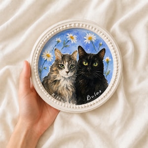 May include: A round white beaded frame containing a watercolor painting of two cats against a blue background with white daisies. The tabby cat on the left is labeled Kitty, and the solid black cat on the right is labeled Bulma in white paint.
