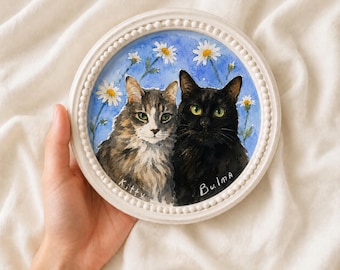Può includere: Una cornice rotonda bianca con perline contenente un acquerello di due gatti su sfondo blu con margherite bianche. Il gatto tigrato a sinistra è etichettato Kitty e il gatto nero a destra è etichettato Bulma in vernice bianca.